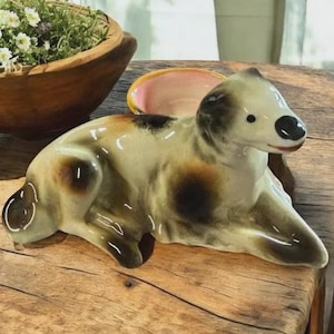 May include: A ceramic dog ornament in a resting pose, with a glossy finish. The dog has a cream-coloured body with brown and black spots. Its eyes and nose are black. The figurine is placed on a wooden surface, with a wooden bowl and a small pink dish in the background.