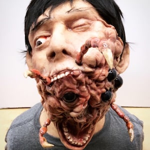 Könnte beinhalten: Eine Nahaufnahme einer realistischen Horror-Maske, die ein menschliches Gesicht mit einem grotesken, fleischigen Wesen zeigt, das aus ihm herauswächst. Das Wesen hat mehrere Augen, scharfe Zähne und Krallen.