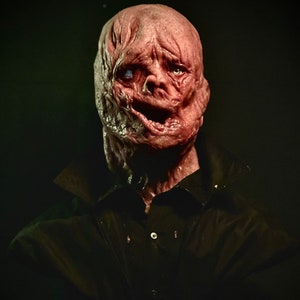 May include: A disturbing, realistic mask with a fleshy, distorted appearance. The mask is predominantly red and pink, with visible wrinkles and a partially open mouth. It is worn over a black collared shirt, creating a horror-themed aesthetic.