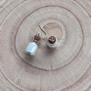 May include: A pair of dangle earrings featuring miniature clear glass bottles filled with tiny iridescent white beads. Each bottle is sealed with a brown cork stopper and attached to a silver-toned metal hook for ear attachment.
