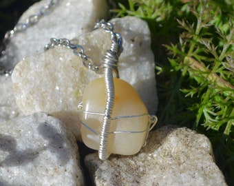 Puede incluir: Un collar de cadena de plata con un colgante de piedra de color marrón claro en forma de lágrima envuelto en alambre. El colgante descansa sobre un lecho de rocas blancas.