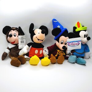 May include: Four Mickey Mouse plush toys in various outfits. One wears a brown winter coat, another a red and black outfit, a third a wizard costume, and the last a blue outfit with a green hat. Each plush has a tag.