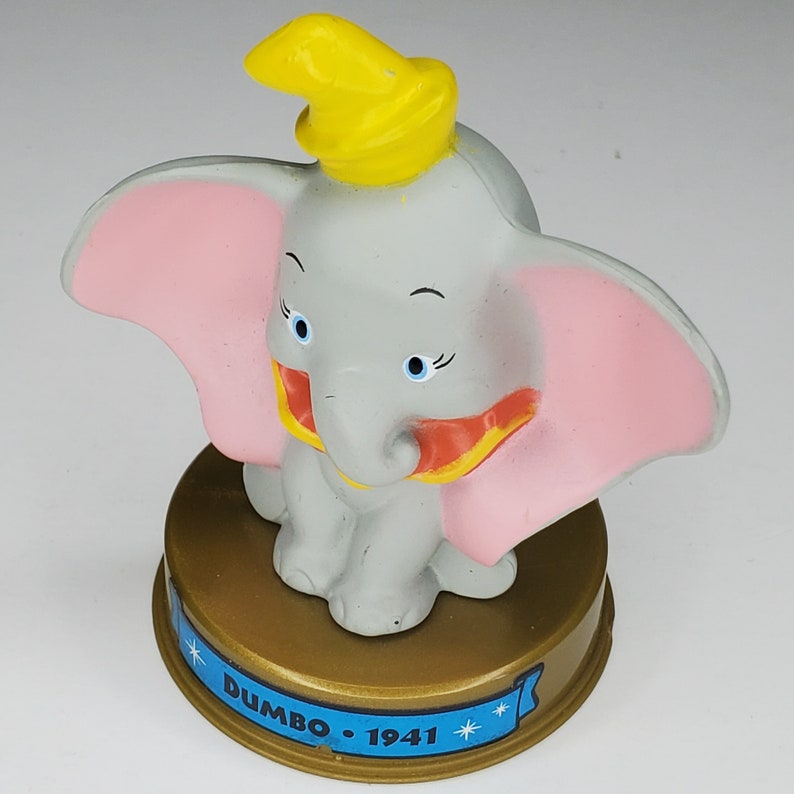 May include: A gray plastic toy figurine of Dumbo the elephant from the Disney movie Dumbo. Dumbo is wearing a yellow hat and a pink cape. The figurine is standing on a gold base with the text "Dumbo - 1941" on it.