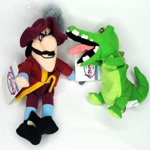 May include: Two plush toys: Captain Hook in a red coat and hat, and a green crocodile with a pink tongue. Both have tags from The Disney Store. The Captain Hook doll has a black mustache and a yellow sash.
