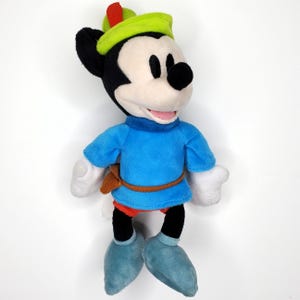 May include: A plush Mickey Mouse toy dressed as Robin Hood. The toy has a green hat with a red feather, a blue tunic, and blue shoes. The toy has black ears, a black nose, and a white face. The toy is approximately 10 inches tall.