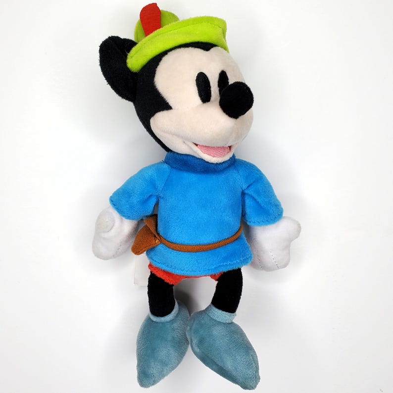 May include: A plush Mickey Mouse toy dressed as Robin Hood. The toy has a green hat with a red feather, a blue tunic, and blue shoes. The toy has black ears, a black nose, and a white face. The toy is approximately 10 inches tall.