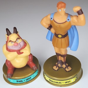 May include: Two plastic figurines of characters from the animated film Hercules. One figure is Hercules, in a brown tunic and blue cape, flexing his arm. The other is Phil, a yellow satyr with red accents. Both figures are on gold bases with the year 1997.