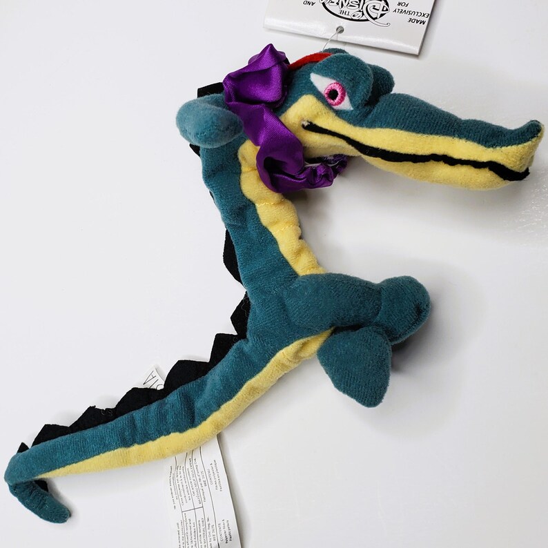 K&ouml;nnte beinhalten: Ein Pl&uuml;schtier-Alligator in T&uuml;rkis, Gelb und Schwarz mit einer lila Schleife. Der Alligator hat eine lange Schnauze, ein rosa Auge und schwarze Stacheln entlang des R&uuml;ckens. Ein Etikett ist am Spielzeug befestigt.