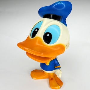 May include: A vintage Donald Duck figurine. The figure has a white head, an orange beak and feet, and a blue sailor hat and jacket. The eyes are black with light blue irises. The figurine is a classic cartoon character.