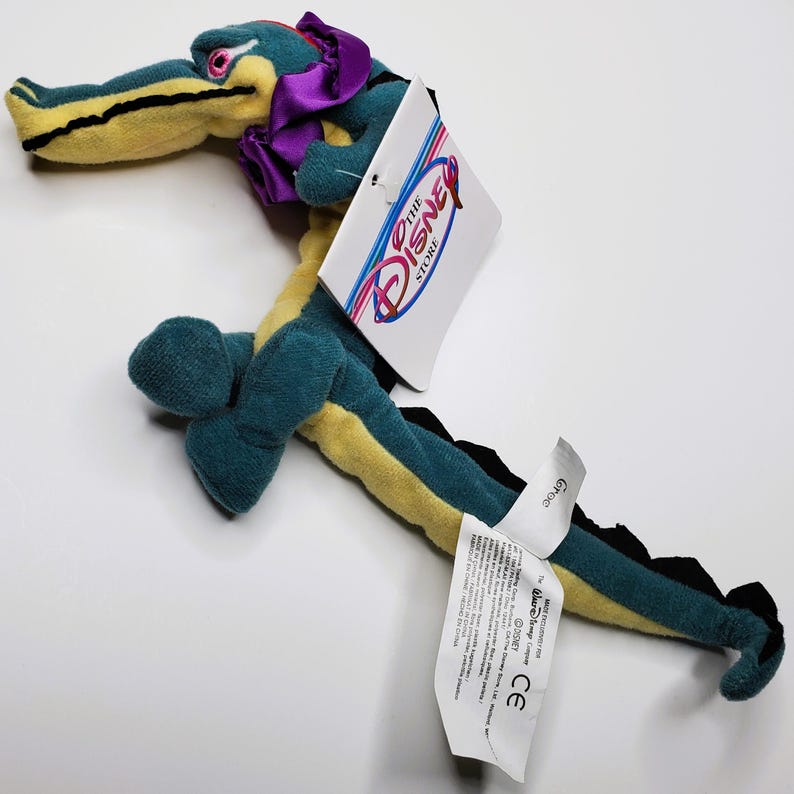 K&ouml;nnte beinhalten: Ein Pl&uuml;schtier-Krokodil in T&uuml;rkis, Gelb und Schwarz, mit einer lila Schleife und einem Disney Store-Etikett. Das Krokodil hat einen langen K&ouml;rper und Schwanz mit detailliertem Design.