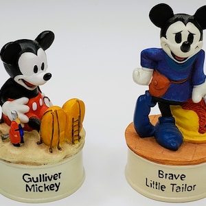 May include: Two Mickey Mouse figurines. One is in a red and black outfit with the text "Gulliver Mickey" on the base. The other is in a blue outfit with the text "Brave Little Tailor" on the base. Both are on cream-colored bases.