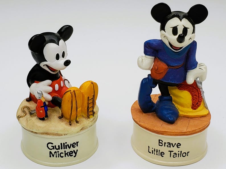 May include: Two Mickey Mouse figurines. One is in a red and black outfit with the text "Gulliver Mickey" on the base. The other is in a blue outfit with the text "Brave Little Tailor" on the base. Both are on cream-colored bases.
