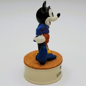 May include: A figurine of Mickey Mouse in a blue jacket, orange pants, and yellow shoes. The black-eared character stands on a round, tan platform atop a cream-colored base. The base has the words "Big Little" printed on it.