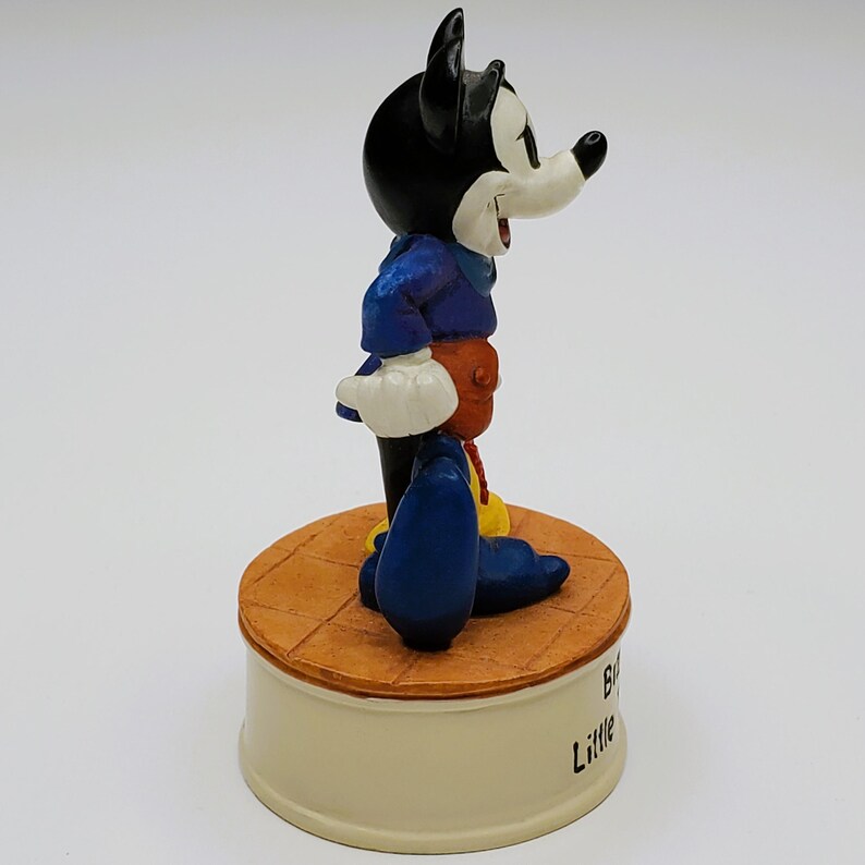 May include: A figurine of Mickey Mouse in a blue jacket, orange pants, and yellow shoes. The black-eared character stands on a round, tan platform atop a cream-colored base. The base has the words "Big Little" printed on it.