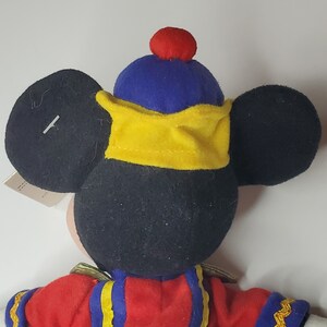 Puede incluir: Un peluche con cabeza negra y grandes orejas redondas, que lleva un gorro azul con un pomp&oacute;n rojo y una banda amarilla. El personaje viste una chaqueta roja con adornos azules y dorados, visible desde la parte posterior y los hombros.