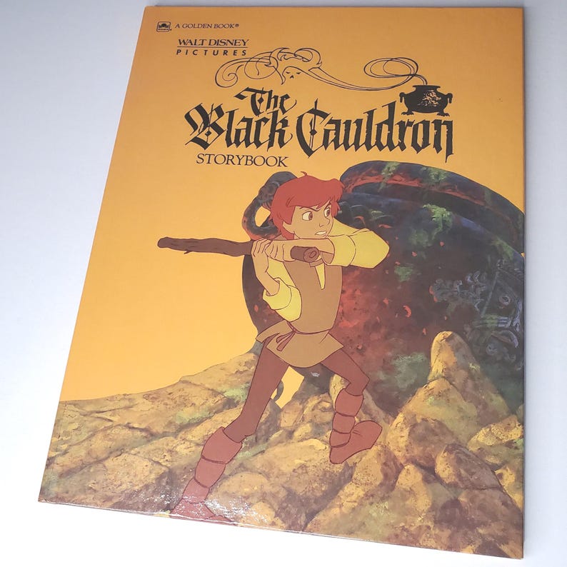 May include: A Golden Book of Walt Disney Pictures' "The Black Cauldron Storybook." The cover features an illustration of a young man with a staff, set against a yellow and brown background with a large cauldron.