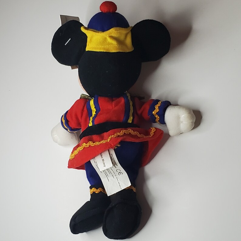 Puede incluir: Un peluche de Mickey Mouse que viste una chaqueta de estilo militar roja y azul con ribetes dorados, un gorro azul con un pomp&oacute;n rojo y una banda amarilla, y botas negras. El juguete tiene guantes blancos y una etiqueta blanca con texto.