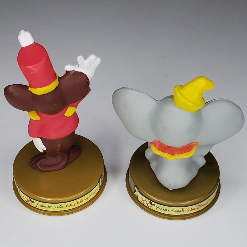 May include: Two plastic figurines, one of a brown and red dressed mouse and the other of a grey elephant, both with yellow hats. They are on gold bases with the text "100 Years of Magic Walt Disney World" on them.