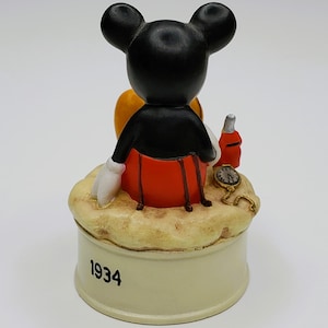 May include: A vintage Mickey Mouse figurine, viewed from the back, with black ears, a black jacket, red shorts, and white gloves. The figurine sits atop a beige base with the year "1934" printed on it. A small bottle and pocket watch are also present.