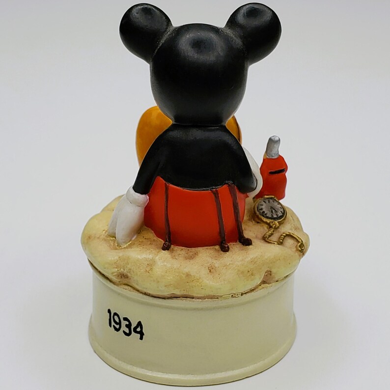 May include: A vintage Mickey Mouse figurine, viewed from the back, with black ears, a black jacket, red shorts, and white gloves. The figurine sits atop a beige base with the year "1934" printed on it. A small bottle and pocket watch are also present.