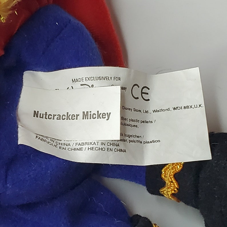 Puede incluir: Un peluche con una etiqueta de tela blanca que muestra el texto Nutcracker Mickey, Made Exclusively For Disney Store y Made In China. El juguete tiene tela azul, una secci&oacute;n roja y tela negra con un borde dorado visible en los extremos.