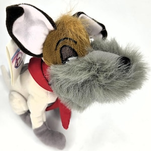 May include: A plush dog toy featuring a tan and white body, large dark brown ears with white inner lining, and a prominent gray shaggy muzzle. The character wears a bright red fabric collar or scarf around its neck and has a small tag attached.