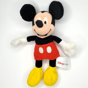 May include: A Mickey Mouse plush toy with a black head and ears, a tan face, white gloves, a red skirt with two white buttons, and yellow shoes. The Disney Store tag is attached.