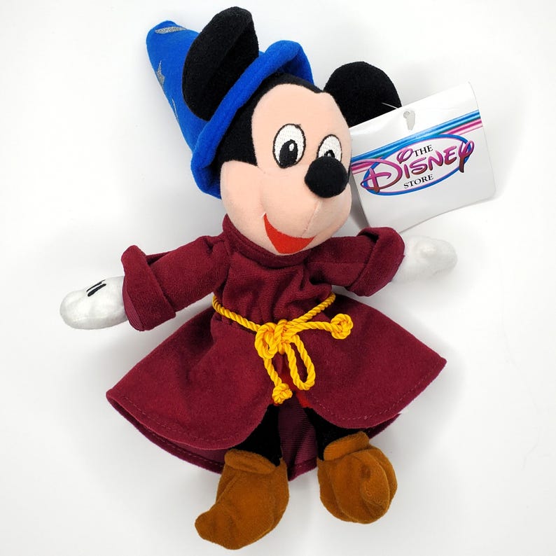 May include: A plush Mickey Mouse toy dressed as a sorcerer, wearing a blue hat with a star, a burgundy robe, and brown boots. The toy has a yellow rope belt and a tag that says "The Disney Store".