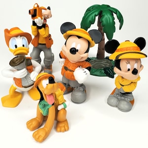 May include: A collection of Disney character figurines in safari outfits. Donald Duck, Goofy, Mickey Mouse, Minnie Mouse, and Pluto are featured, each wearing a safari hat and clothing. A green palm tree is also included in the display.