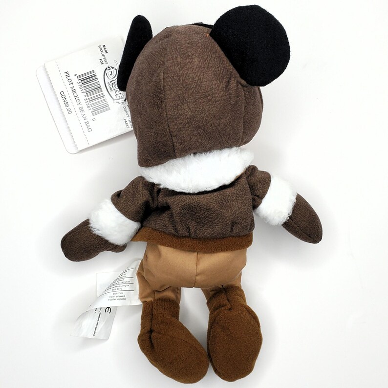 May include: A plush Mickey Mouse Pilot Bean Bag doll, viewed from the back. The doll is dressed in a brown aviator jacket and hat with white trim. The doll has black ears and brown pants. A tag is attached with the text "Pilot Mickey Bean Bag".