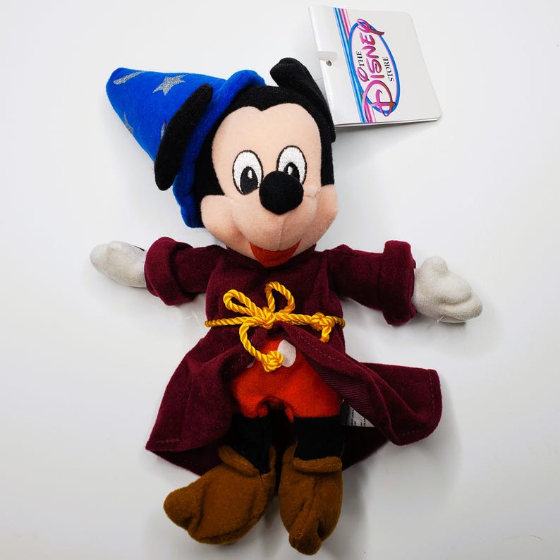 K&ouml;nnte beinhalten: Ein Mickey Mouse Pl&uuml;schtier als Zauberer verkleidet, mit einem blauen Hut mit silbernen Sternen, einer weinroten Robe und braunen Schuhen. Das Spielzeug hat einen gelben Seilg&uuml;rtel und wei&szlig;e Handschuhe. Ein Disney Store-Etikett ist angebracht.