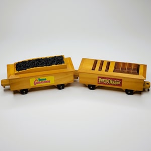 May include: Two wooden toy train cars. One car is filled with black material, and the other has wooden accents. The first car has a yellow label with the text "The Three Caballeros". The second car has a red label with the text "Pete's Dragon".