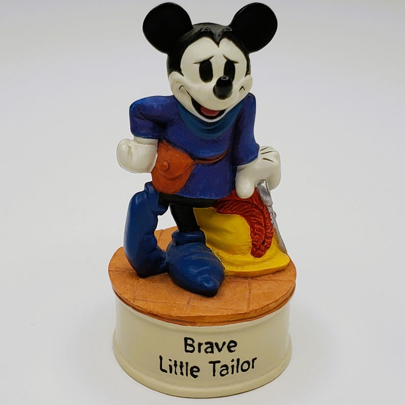 May include: A ceramic figurine of Mickey Mouse in a blue outfit, holding a sword and leaning on a yellow sack. The figurine is on a round base with the words "Brave Little Tailor".