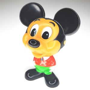 May include: A vintage Mickey Mouse figurine. The figure has a black head and ears, a yellow face, and a red shirt with green pants. The figurine is standing with its arms at its sides.