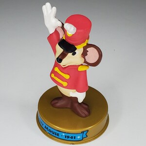 May include: A red and gold plastic figurine of Mickey Mouse dressed as a drum major. He is wearing a red uniform with gold trim and a red hat with a gold band. The figurine is standing on a gold base with the words "Mickey Mouse 1941" printed on it.