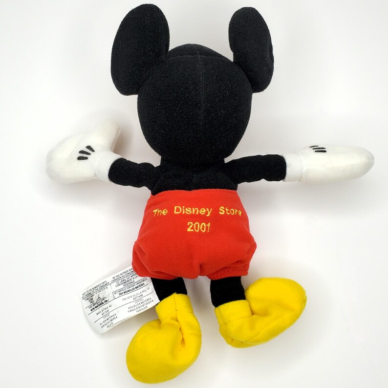 May include: A Mickey Mouse plush toy with black ears, head, and arms, white gloves, red shorts, and yellow shoes. The Disney Store 2001 is embroidered on the back. A white tag is attached to the toy.