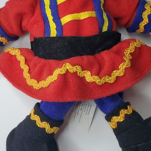 Puede incluir: Una mu&ntilde;eca de peluche con una parte superior de fieltro rojo con detalles de cintas azules y amarillas, una cintura negra y una falda roja adornada con cinta de zigzag dorada. La mu&ntilde;eca tiene piernas azules y botas de fieltro negro con borde dorado.