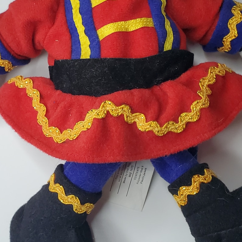Puede incluir: Una mu&ntilde;eca de peluche con una parte superior de fieltro rojo con detalles de cintas azules y amarillas, una cintura negra y una falda roja adornada con cinta de zigzag dorada. La mu&ntilde;eca tiene piernas azules y botas de fieltro negro con borde dorado.