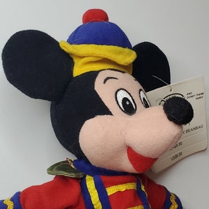 Puede incluir: Un peluche de Mickey Mouse tipo beanbag que viste una chaqueta roja con ribetes dorados y un gorro azul y amarillo con un pomp&oacute;n rojo. El juguete tiene orejas negras, cara color melocot&oacute;n y una etiqueta blanca que dice Disney Theme Parks.