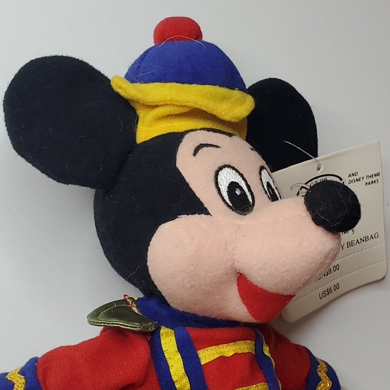 Puede incluir: Un peluche de Mickey Mouse tipo beanbag que viste una chaqueta roja con ribetes dorados y un gorro azul y amarillo con un pomp&oacute;n rojo. El juguete tiene orejas negras, cara color melocot&oacute;n y una etiqueta blanca que dice Disney Theme Parks.