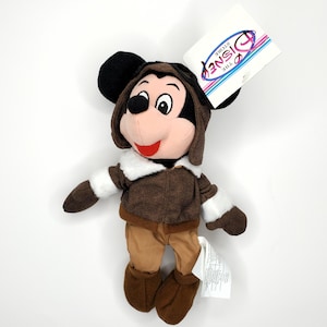 May include: A Mickey Mouse plush toy wearing a brown aviator jacket, pants, and boots, with a matching cap. The plush has black ears, a pink face, and a red smile. A Disney Store tag is attached.