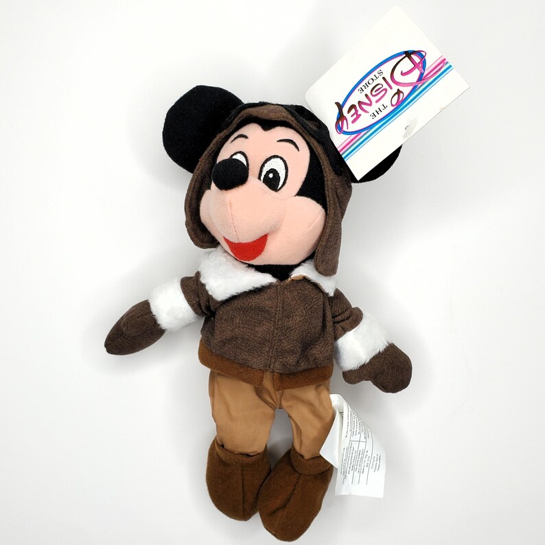 May include: A Mickey Mouse plush toy wearing a brown aviator jacket, pants, and boots, with a matching cap. The plush has black ears, a pink face, and a red smile. A Disney Store tag is attached.