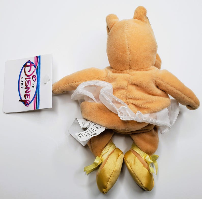 K&ouml;nnte beinhalten: Ein Pl&uuml;schtier einer Figur aus Disneys Fantasia, mit einem wei&szlig;en Tutu und goldenen Ballettschuhen. Das Spielzeug ist hellbraun und hat ein Disney Store-Etikett. Die Figur hat kleine Ohren und ausgestreckte Arme.