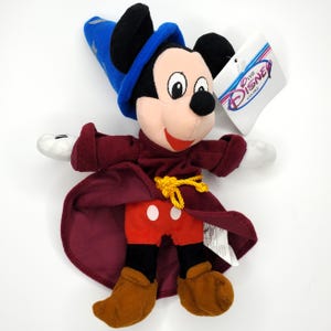 May include: A plush Mickey Mouse dressed as a sorcerer, wearing a blue hat with stars, a burgundy robe, and red shorts with white polka dots. The toy has black ears, a black nose, and brown shoes. A tag from The Disney Store is attached.