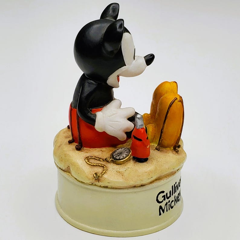 May include: A Mickey Mouse figurine in a seated pose, wearing red shorts and white gloves. The figurine is on a sand-colored base with a pocket watch and a red bottle. The base has the words "Gulliver Mickey".