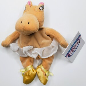 K&ouml;nnte beinhalten: Ein beiges Pl&uuml;schpferd mit einem wei&szlig;en Tutu und goldenen Ballettschuhen. Das Nilpferd hat rosa Akzente an den Ohren. Ein Etikett von The Disney Store ist angebracht.