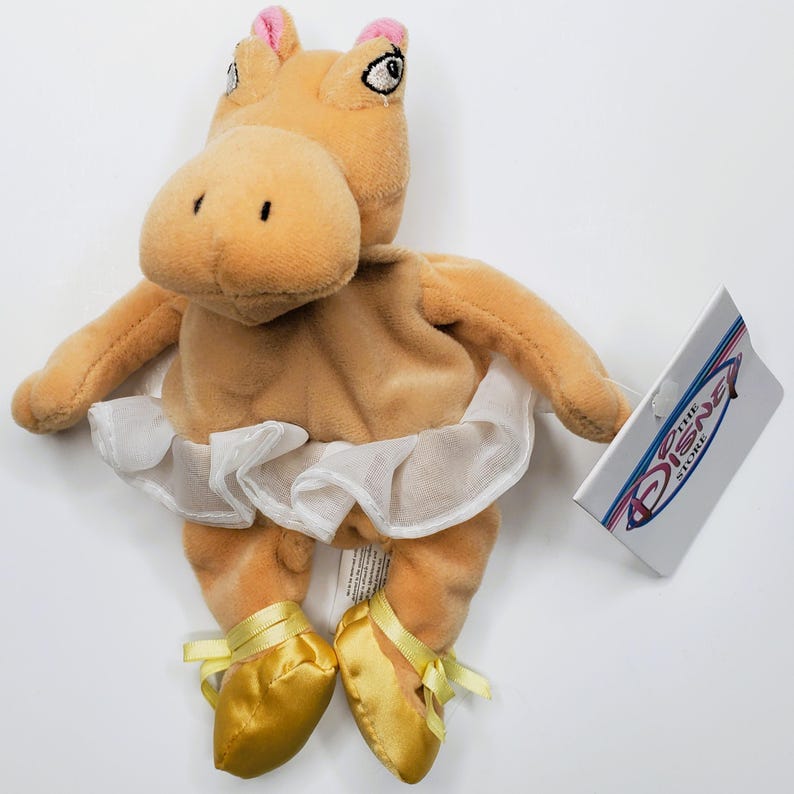 K&ouml;nnte beinhalten: Ein beiges Pl&uuml;schpferd mit einem wei&szlig;en Tutu und goldenen Ballettschuhen. Das Nilpferd hat rosa Akzente an den Ohren. Ein Etikett von The Disney Store ist angebracht.
