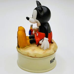 May include: A vintage Mickey Mouse figurine, featuring the iconic character in black, red, and white, seated on a sandy base. The figurine is resting on a cream-colored base with the year "1934" inscribed. A collectible Disney item.