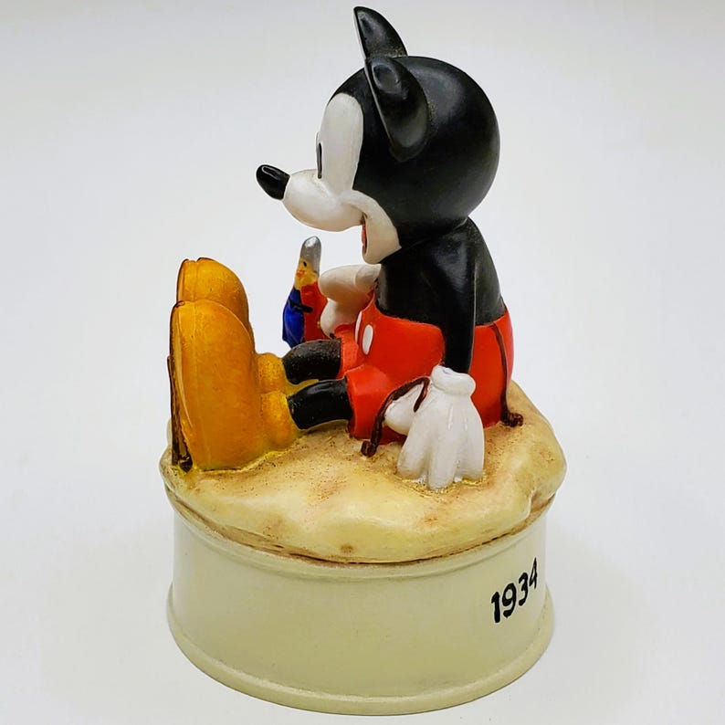 May include: A vintage Mickey Mouse figurine, featuring the iconic character in black, red, and white, seated on a sandy base. The figurine is resting on a cream-colored base with the year "1934" inscribed. A collectible Disney item.