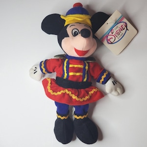 Puede incluir: Un mu&ntilde;eco de peluche de Mickey Mouse con un uniforme rojo y azul con adornos dorados, un sombrero a juego y guantes blancos. El mu&ntilde;eco tiene una cara sonriente y una etiqueta de color crema con el texto The Disney Store en azul y rojo.